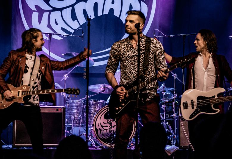 Kuvassa on Aston Kalmarin kolme soittajaa esiintymässä, joista keskellä on laulaja ja molemmin puolin on kaksi kitaristia.  Kuvan sävy on tumma ja valot violetin sävyisiä.  Taustalla on bändin nimi isolla taustaseinällä.