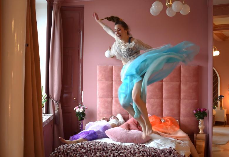 Kuvassa on  on tanssija hyppäämässä sängyn päällä roosan värisessä huoneessa ja taustalla näkyy toista huonetta. Tanssijalla on vaalean sininen tyllihame ja vaalea pusero.