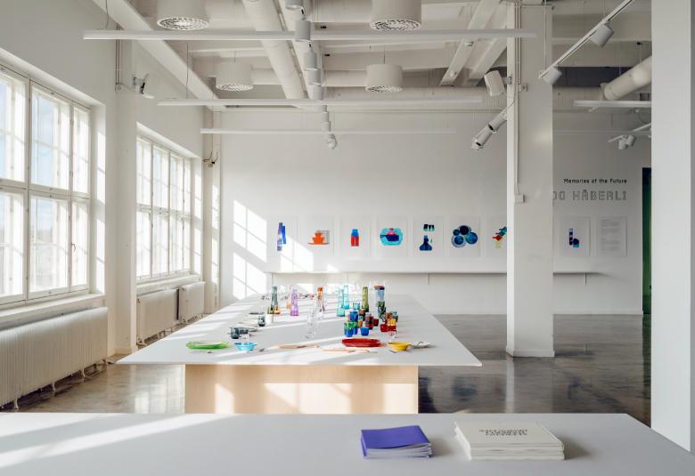 Kuvassa on näyttelytilaa ja iso valkoinen pöytä, jossa on taide-esineitä. Vasemmalla on iso ruudutettu ikkuna.