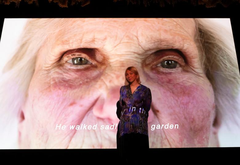 Kuvassa on Emilija Škarnulytė seisomassa suuren teoksensa edessä. Takana on suuri kasvokuva hänen sokeasta isoäidistään. Kuvassa on mustat reunukset.