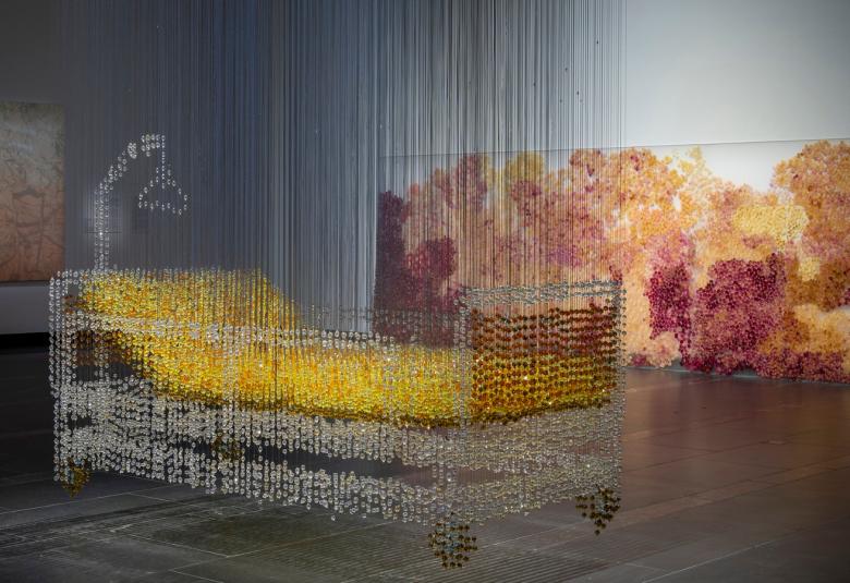 Kuvassa on keskellä installaationa sänky, jossa lasisen näköiset laidat ja pohja sekä päädyssä nousutuki. Patja on keltainen.  Taustalla on tauluja.