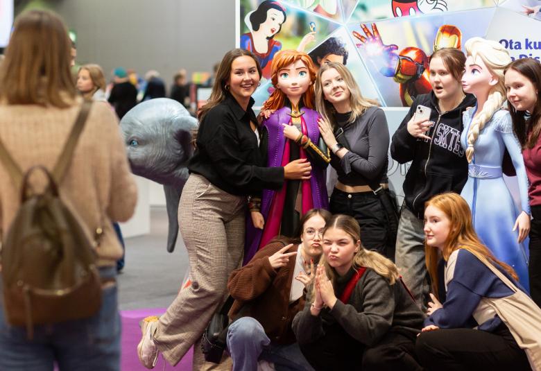 Kuvassa on nuoria naisia ryhmäkuvassa ilmeikkäinä ja kaksi nuorta naista pitää keskellään sarjakuvahahmonukkea.  Taustalla on Disneyn kirjajuliste.