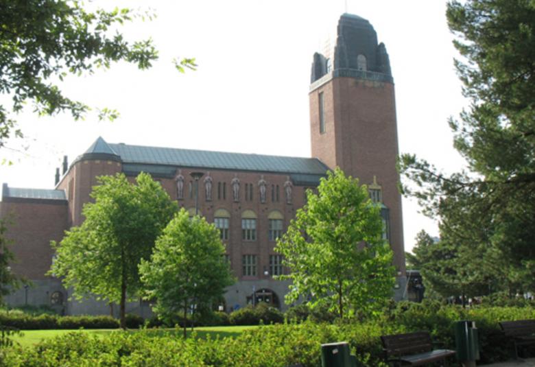 Kuvassa on Joensuun kaupungintalo ja se on punatiilinen kookas rakennus. Sen edustalla on vihreitä. lehtipuita ja pieni puisto.