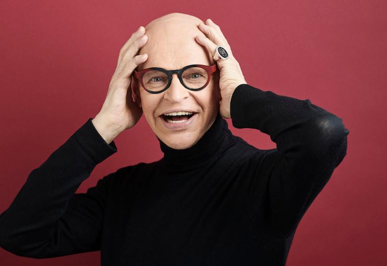 Kuvassa on Jorma Uotinen ja hän pitää molempia käsiään päänsä sivuilla. Hänellä on musta poolopaita päällään ja tausta on punertavan ruskea.