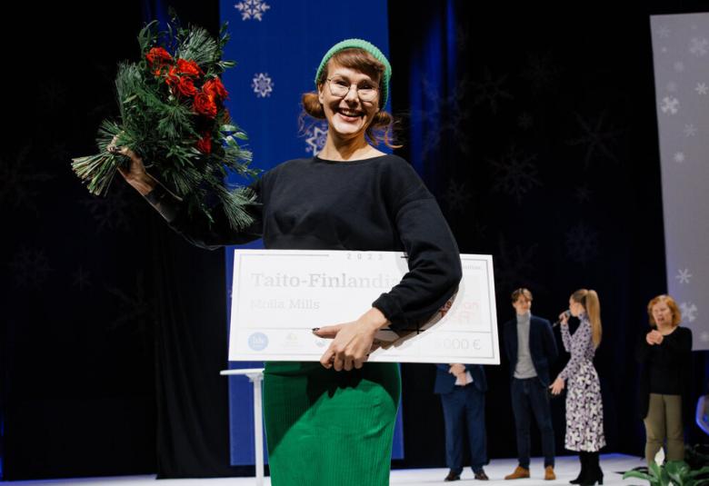 Kuvassa on Molla Mills ja hänellä on oikeassa kädessään ruusukimppu. Hänellä on päällään mursta t-paita ja vihreät housut. Taustalla sinistä seinämää ja oikealla on taustalla muutamia henkilöitä seisomassa kauempana. Toisessa kädessä on iso kunniakirja.