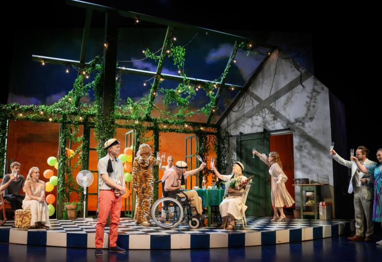 Kuvassa on näyttämö, jossa on kahdeksan näyttelijää juhlimassa vappua.  Näyttämöllä on rakennelma, jossa on vihreitä köynnöksiä. Etualalla on nuori mies teekkarilakki päässään.