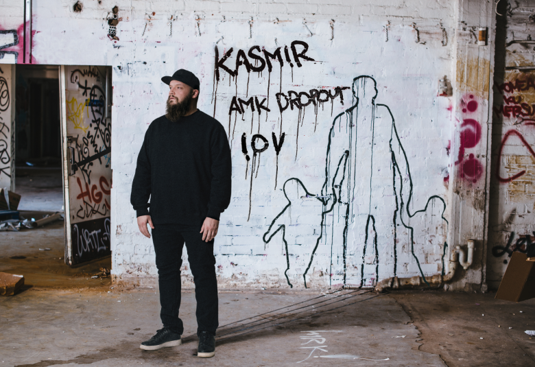 Kuvassa on Kasmir seisomassa  betoniseinien keskellä ja hänen takanaan lukee vaalealla seinällä lukee Kasmir AMK Dropout 10 v. Vieressä on ääriviivakuvat miehestä, joka pitää kädestä kiinni kahta lasta.