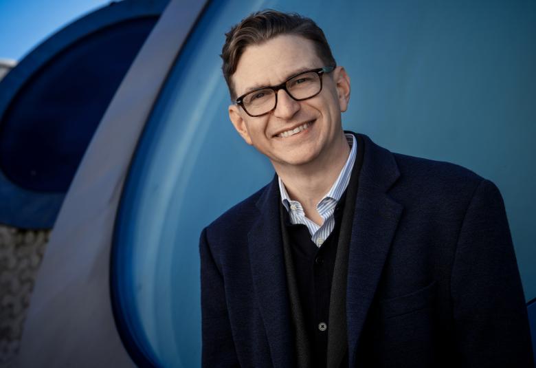 Kuvassa on Kieran Long puolivartalokuvassa Amos Rexin pihalla ja hänellä on musta päällystakki päällään. Taustalla on tummia kaaria, jotka ovat Amos Rexin pihan kumpuja.