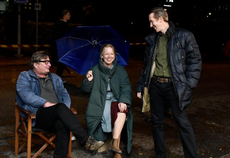 Kuvassa ovat pimeässä istumassa tuoleilla Aki Kaurismäki, Alma Pöysti ja seisomassa Jussi Vatanen. He juttelevat.