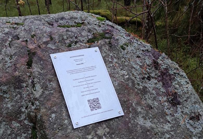 Kuvassa on iso kivi metsässä  ja siihen on kiinnitetty A4-kokoinen, metallinen laatta. Siinä on otsikko Kuunteluasentoja ja ohjetekstiä muutamalla rivillä ja QR-koodi.