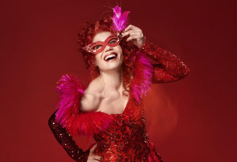 Kuvassa on Helena Juntunen, jolla on punainen avonainen puuhkamainen avara kaula-aukko ja kiiltävät hihat. Hiukset ovat myös punaiset sekä kuvan tausta. Kuva on puolivartalokuva.