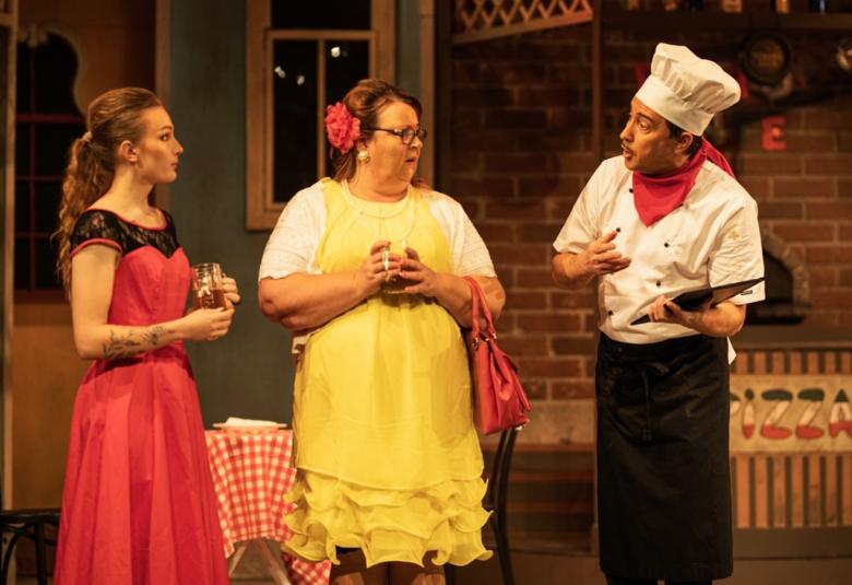 Kuvassa on kaksi naista seisoo keskustelemassa pizzakokin kanssa. Vasemman puoleisella naisella on punainen mekko ja hänen vieressään olevalla keltainen mekko päällään.  Tila on pizzaravintola.