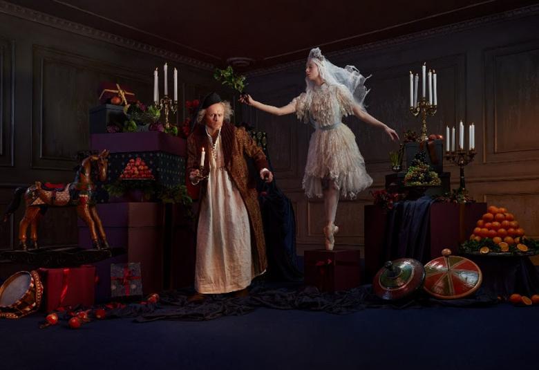 Kuvassa ovat tummassa huoneessa saituri Scrooge ja hänestä vinosti yläpuolella ballerina valkoisessa puvussa. Huoneessa on hedelmiä ja kynttilöitä.