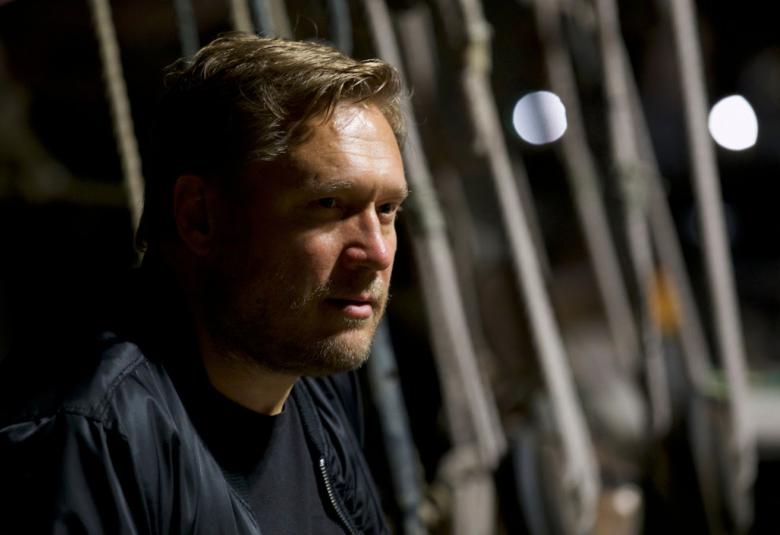 Kuvassa on Samuli Edelmann puolivartalokuvassa istumassa sivuttain ja valoa näkyy puolittain hänen kasvoillaan. Taustalla näkyy gunneöstu teräsputkia pystysuorassa.