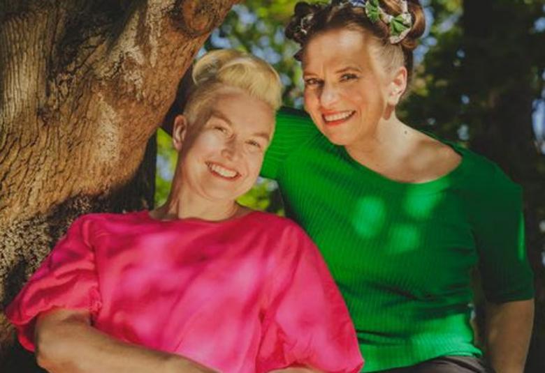 Kuvassa ovat Satu Rämö ja Satu Kontinen isoon puun runkoon nojaamassa. Rämöllä on pinkin punainen ja Kontisella kirkkaan vihreä pusero päällään.