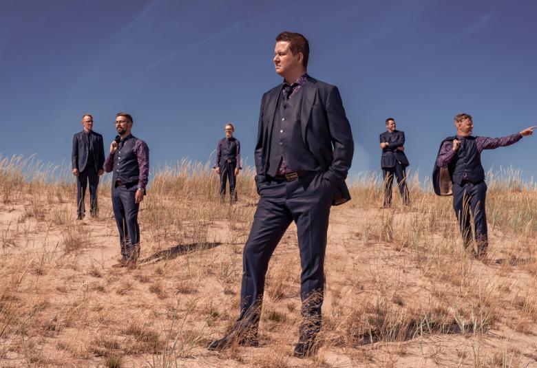 Kuvassa on Sinitaivaan kuusi jäsentä seisomassa hiekkarannalla, jossa harvaa heinikkoa. Yksi jäsenistä on etummaisena ja kaikilla on puvut päällään. Taustalla on kesäistä taivasta.