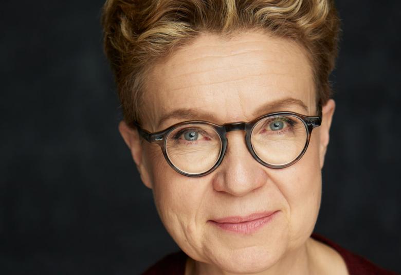 Kuvassa on Sirpa Kähkönen kasvokuvassa. Hänellä on ruskeat, lyhyet hiukset ja mustakehyksiset silmälasit. Tausta on tumma.