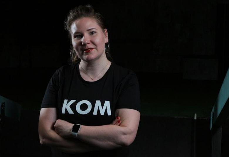 Kuvassa on Susanna Airaksinen ja hän on puolivartalokuvassa. Tausta on tumma. Hänellä on musta t-paita, jossa lukee KOM.