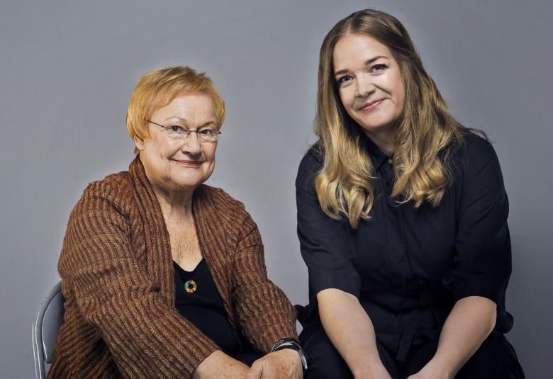 Kuvassa ovat istumassa Tarja Halonen ja Meri Valkama. Halosella on ruskea takki päällään ja Valkamalla on musta mekko. Tausta on harmaa.