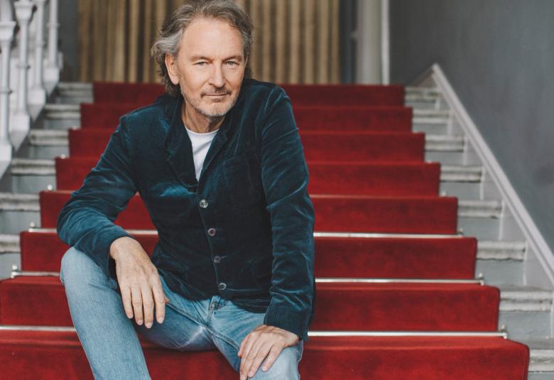 Kuvassa on Tomas Ledin istumassa rennosti sisätilassa olevilla rappusilla.  Niillä on leveä punainen matto harmailla portailla. Kaiteissä on harmaat koristeelliset kaidetolpat.