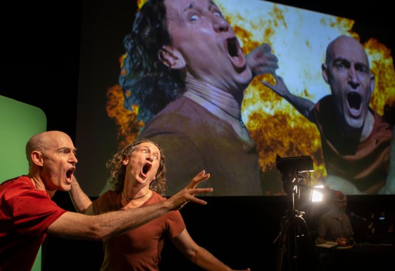 Kuvassa ovat The Umbical Brothersin kaksi miesesiityjää ja he ovat vinottain kädet ojennettuina ja heillä on ruskean punaiset t-painat. Taustalla on iso näyttö, joissa näkyvät heidän ilmeensä.