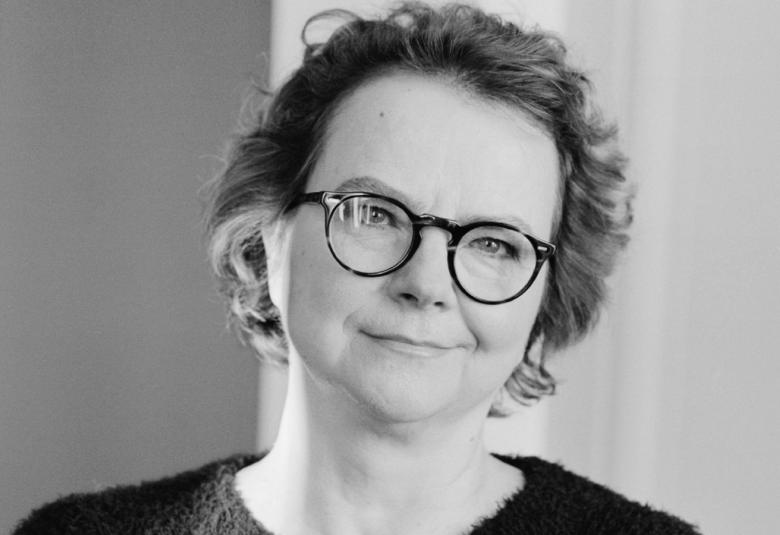 Kuvassa on Laura Kolbe puolivartalokuvassa.  Hänellä on lyhyehköt kiharat hiukset ja silmälasit. Hänellä on päällään tumma villapaita. Kuva on musta-valkoinen. 