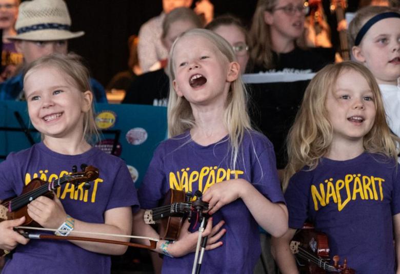 Kuvassa on istumassa kolme tyttöä, joilla on siniset t-paidat ja niissä lukee keltaisella isoin kirjaimin Näppärit. Heillä on soittimet kädessään.