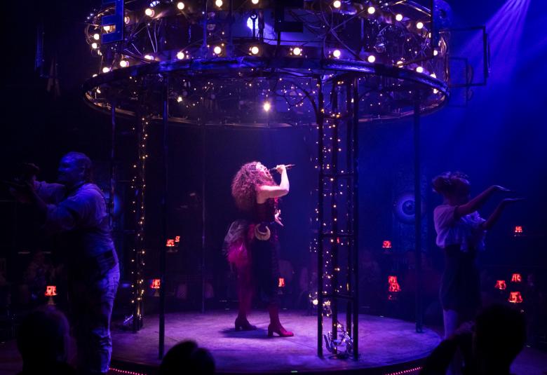 Kuvassa on karusellin keskellä laulamassa Anen Syysmaa ja hän on sivuttain.  Kuvan sävy on tumma ja lilan värinen valaistus. Ympärillä pimeässä näkyy esiintyjiä eri suunnissa.