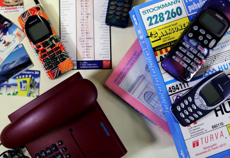 Kuvassa on mustia, pieniä matkapuhelimia pöydän päällä ja alhaalla on viininpunainen pöytäpuhelin, jossa on näppäimet.  Lisäksi on postikortteja ja luottokortteja. Kahden puhelimen alla on oikealla kaksi puhelinluetteloa.