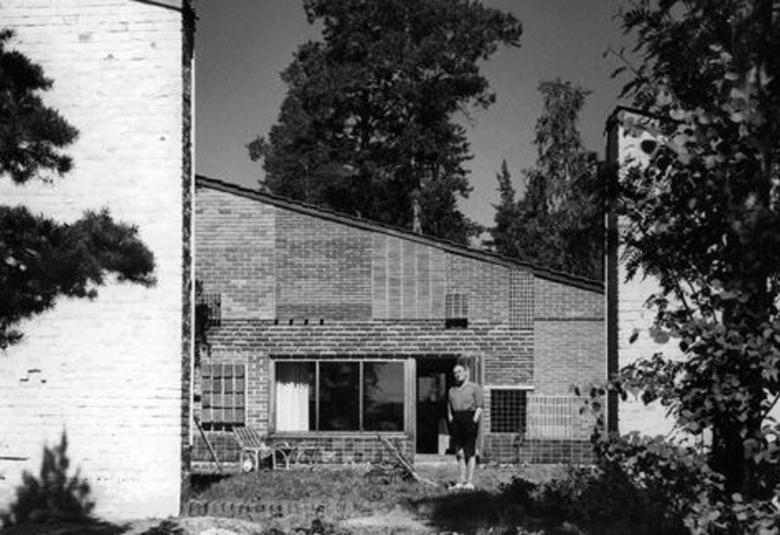 Kuvassa on talo, jossa on vaalea tiiliseinä vasemmalla ja tummempaa talon seinää.  Siinä on vino katto ja siinä on kolmijakoinen ikkuna. Oikealla on vaaleaa tiiliseinää puun takana.  Talon edessä seisoo mies.