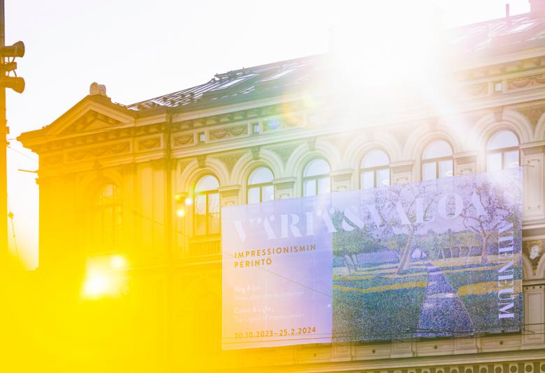 Kuvassa on Ateneumin päärakennuksen julkisivu ja siihen osuu keltainen valo vasemmalta ja oikealta tulee auringon valoa.  Seinällä on iso mainoslakana.