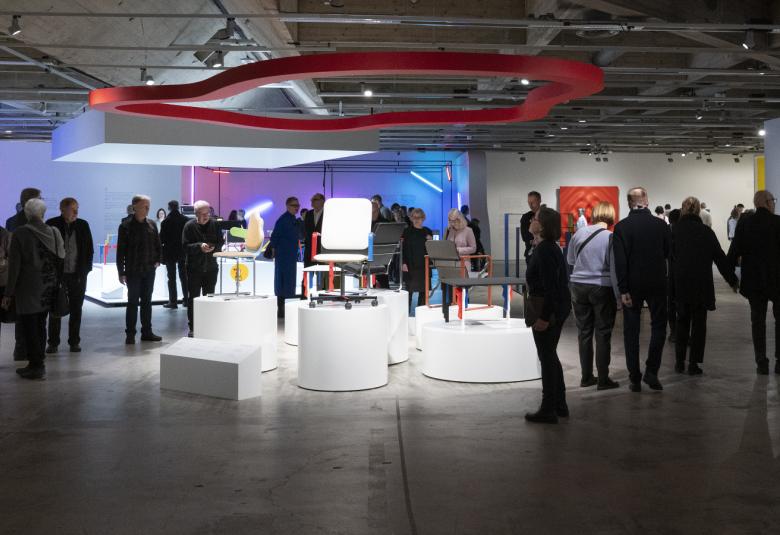 Kuvassa on museokävijöitä vasemmalla sekä oikealla ja keskellä on valkoisia pyöreitä ja neliskulmaisia istuimia.   Tilan on matala ja siellä on valkoisia ja punainen putkielementti.  