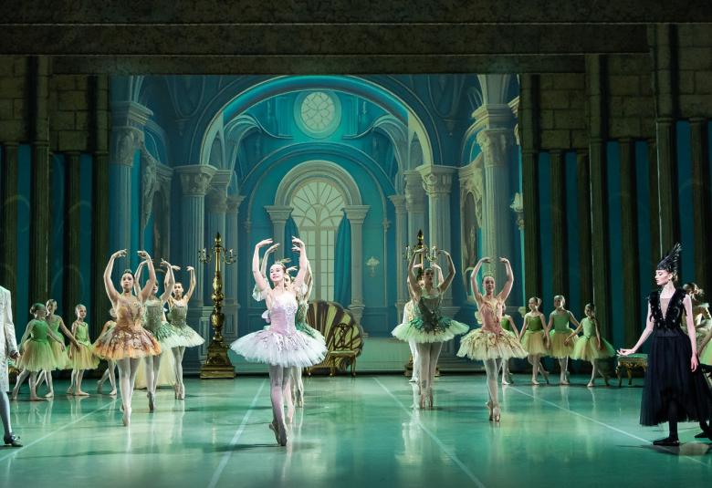  Kuvassa on kansallisbaletin näyttämö ja siinä ovat eri-ikäiset balettikoululaiset ovat tanssimassa lavalla.  Keskellä ovat varttuneet opiskelijat tanssimassa prinsessa-asuissa.  Tausta ja lattia ovat turkoosin värisiä.