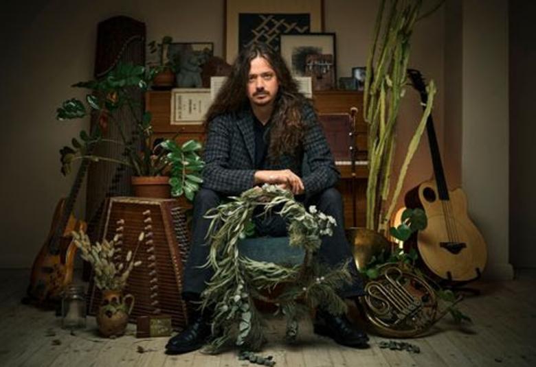 Kuvassa on Lauri Purra istumassa keskellä kuvaa. Taustalla on piano ja sivulla eri ja sivuilla muita soittimia sekä viherkasveja.  Tausta on tumma.