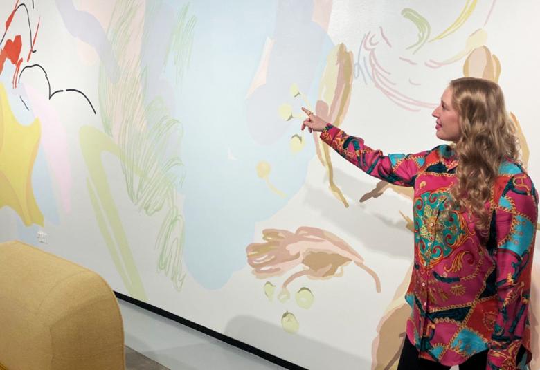 Kuvassa on oikealla Jenni Niskala seinämaalauksen edessä ja hän osoittaa oikealla kadellään maalausta. Hänellä on punaisella ja turkoosilla kuvioitu mekko. Seinämaalaus on vaalean hennon värinen  ja siinä on vaalean sinistä ja vihreää.  Pinnassa on keltainen tähtimäinen kuvio, mustia eri muotoisia viivoja.  