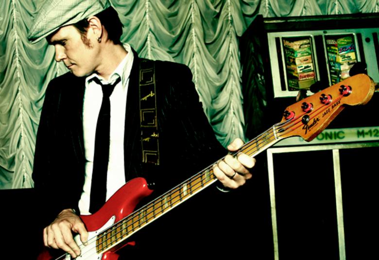 Kuvassa on Marko Haavisto mustassa takissa ja katso vasemmalle alas.  Hänellä on vaalean vihreä lippalakki. Hän soittaa puna.valkoista kitaraa.  Oikealla on tumman ruskea radio.