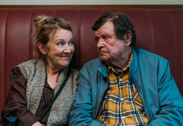 Kuvassa ovat Jaana Saarinen vasemmalla ja Heikki Kinnunen oikealla istumassa tumman violetilla sohvalla, josta näkyy korkea selkänoja. Saarisella on harmaa liivi ja violetti paita.  Kinnusella on sininen pusakka ja kelta-siniraidallinen paita.  Kuva on puolivartalokuva.