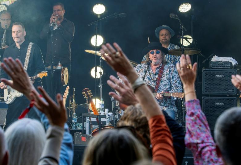 Kuvassa on Hector esiintymässä Salmelan lavalla ja taustalla on bändiä, jossa on kitaristi ja puhaltajia soittamassa.  Edessä on yleisöä kädet pystyssä ja näkyy päälakia. 