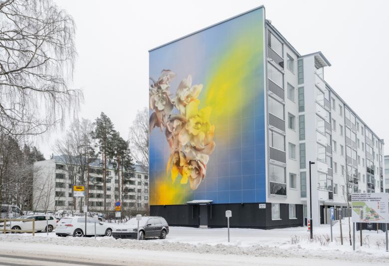 Kuvassa on talvista kerrostaloaluetta ja siinä on vinosti lähin iso kerrostalo. Sen päädyssä on vasemmalta alaspäin lähtevä omenapuun kukan oksa.  Vasemmalta alhaalta ylöspäin nousee keltaista savua.  Tausta on taivaan sininen.