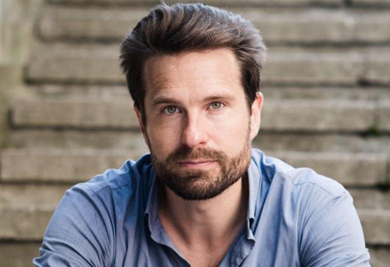 Kuvassa on Valtteri Raekallio, joka istuu taustalla näkyvillä betoniportailla. Hänellä on tummaa partaa ja päällään vaalean sininen paita.