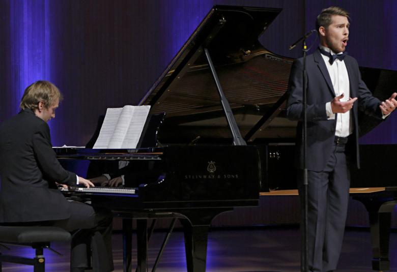 Kuvassa on sivuttain pianisti Juho Alakärppä flyygelin ääressä soittamassa.  Lavalla oikealla on laulamassa Aarne Pelkonen. Taustalla on sini-mustaa verhoa.