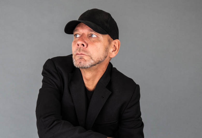 Kuvassa on Kenneth Kvarnström istumassa hiukan vinottain ja katse oikealle. Hänellä on mustat vaatteet ja musta lippalakki päässään.  Kuvan on puolivartalokuva.  Tausta on harmaa.