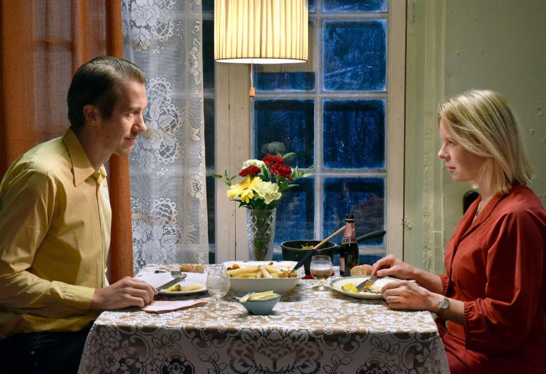 Kuvassa ovat vasemmalla Jussi Vatanen ja oikealla Alma Pöysti istumassa pöydän ääressä ruokailemassa. Taustalla on ruskeat verhot ja valkoiset pitsiverhot sekä ruutuikkuna.