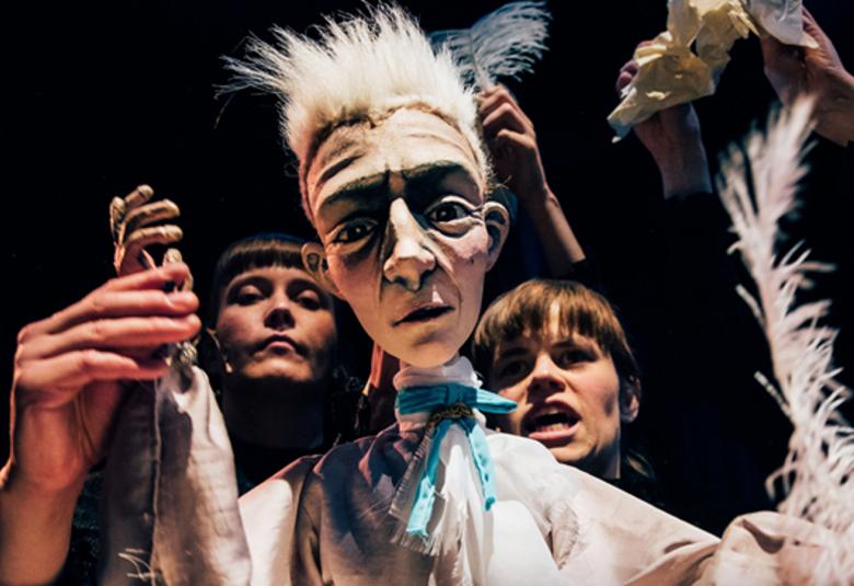 Kuvassa on nukketeatterihahmo, joka on nuori mies ja sillä on vaalea harjastukka.  Vasenta kättä pitelee taustalla nuori näinen ja toisella puolella on nuori mies.  Tausta on tumma.