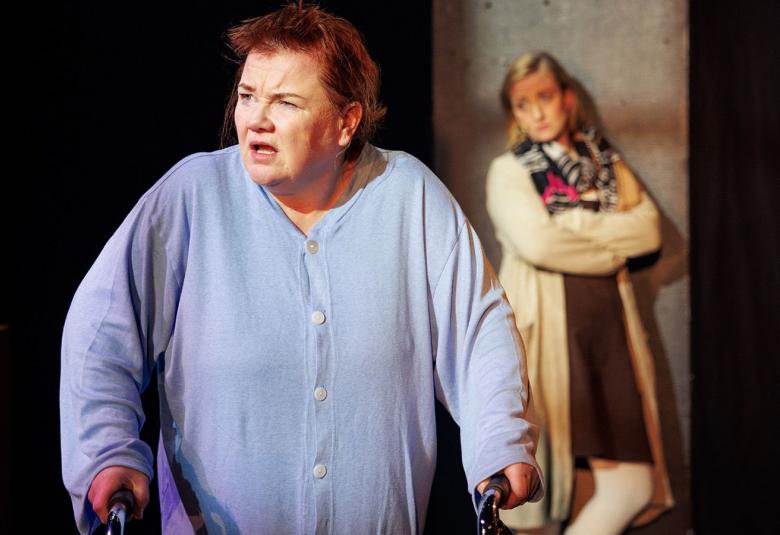 Kuvassa on Minna Kivelä vaalean sinisessä sairaalan paidassa ja rolaattorin kanssa.  Taustalla näkyy nuorempi nainen nojaamassa seinään pitkässä vaaleassa villatakissa.