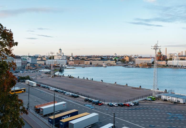 Kuvassa on Helsingin eteläsataman aluetta ja siinä näkyy tontti, jolle arkkitehtuuri- ja designmuseo tulee. Kuvassa näkyy kaukana tuomiokirkko ja kauppatoria sekä etualalla rekkoja parkissa. Taivas on sininen.
