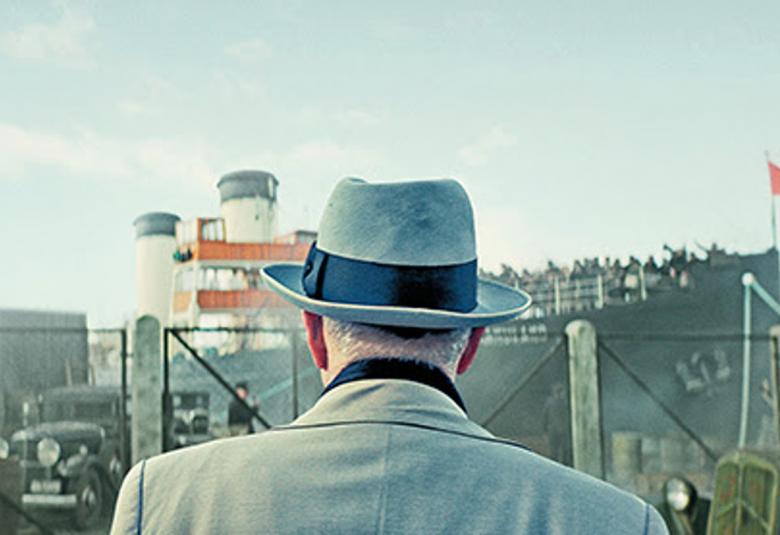 Kuvassa on mies takaapäin siten, että näkyy harmaata takkia ja hattu. Edessä näkuu satamassa olevaa laivaa ja sinistä taivasta.