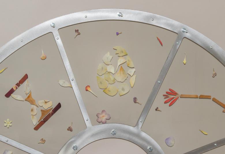 Kuvassa on harmaalla pohjalla teräksen väristä pyörää, joka on jaettu osiin.  Niissä on kultaisen värisiä lehtiä ja vasemmalla on tiimalasi. Oikealla on ruskea litteä kukka.