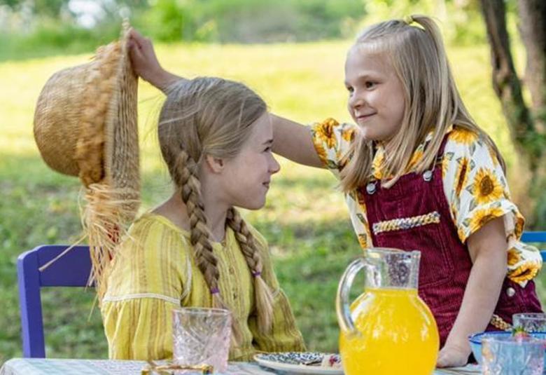 Kuvassa ovat Heinähattu ja Vilttitossu ja Vilttitossu ottaa hattua Heinähatun päästä. He ovat nurmikolla pöydän ääressä, jossa on mehukannussa keltaista mehua.