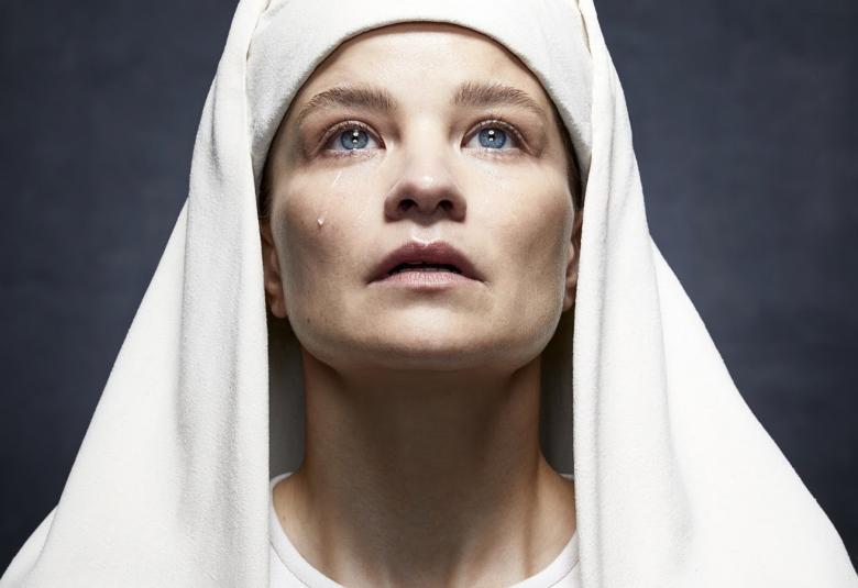 Kuvassa on ylöspäin katsovat naisen kasvot valkoisessa nunnapäähineessä. Tausta on musta.